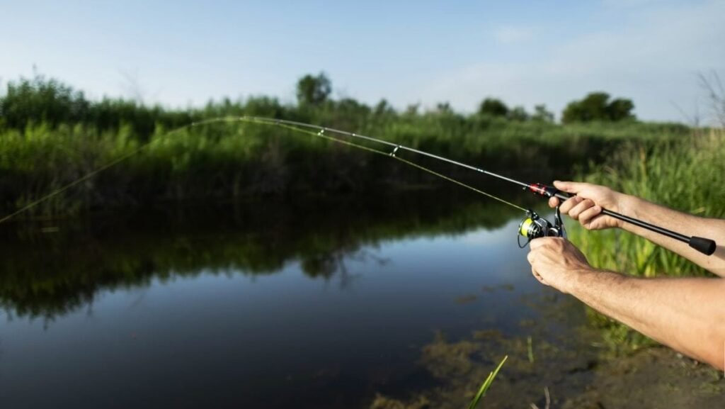 Un bărbat care prinde pește trăge de undiță în timp ce pescuiește dintr-un lac. Pescarul are în mână o undiță cu mulinetă și stă pe malul râului, unde se vede un răsărit de soare.