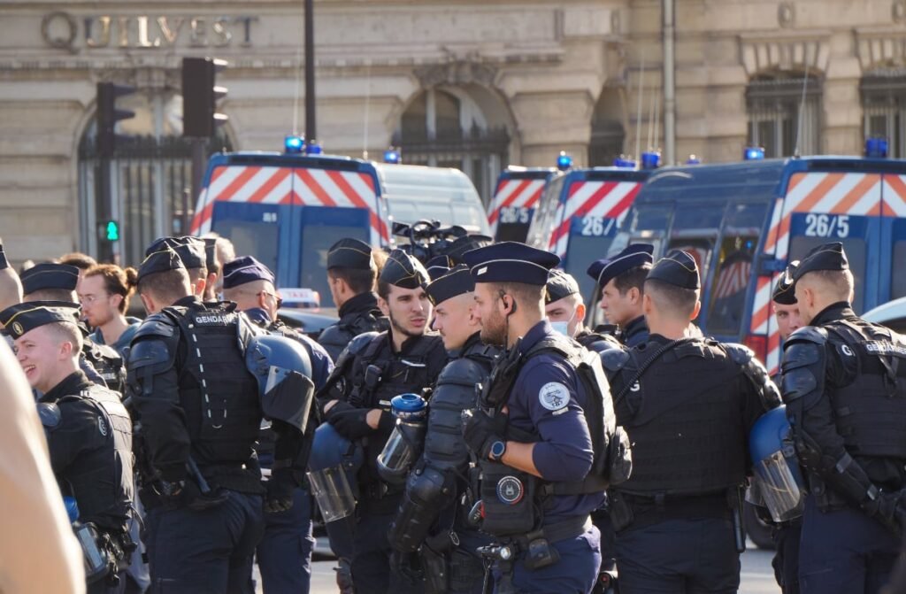 Imaginea arată un grup de ofițeri de poliție și jandarmi îmbrăcați în uniforme de culoare închisă, cu veste de protecție și căști. Unii dintre ei poartă berete negre, iar unii par să aibă părul tuns scurt. O parte din ofițeri stau de vorbă între ei. În fundal se pot vedea mai multe dube de poliție albastre, cu dungi roșii și albe, sugerând că se află în timpul unei misiuni sau că patrulează.