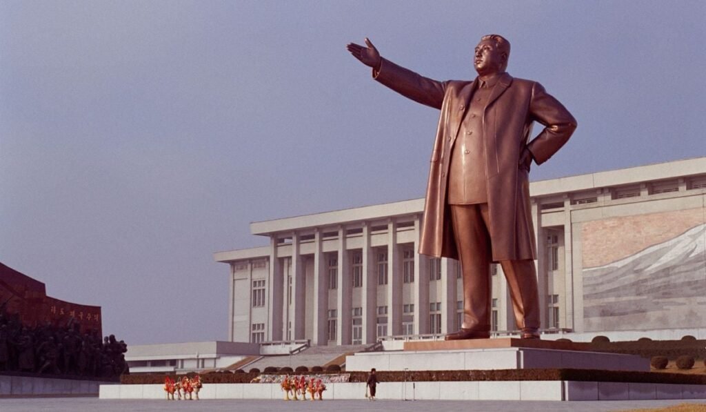 monument înalt, din material de culoarea bronzului, care îl înfățișează pe liderul nord-coreean Kim Jong-Un, cu o mână în șold și cealaltă mână întinsă în față, în fața unei clădiri albe cu coloane, Pyongyang, Coreea de Nord