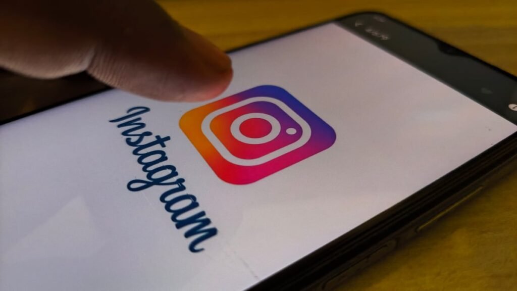 Imaginea prezintă un prim-plan al ecranului unui smartphone pe care este afișată sigla și numele aplicației Instagram. Un deget se apropie sau atinge pictograma, sugerând momentul deschiderii sau interacțiunii cu aplicația.