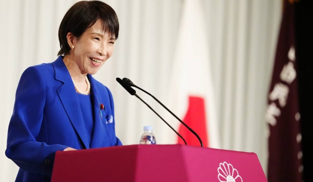 Sane Takaichi, prima femeie premier din istoria Japoniei, îmbrăcată într-un costum albastru, în timp ce vorbește la microfon la o conferință de presă, zâmbește și are buzele date cu ruj roșu