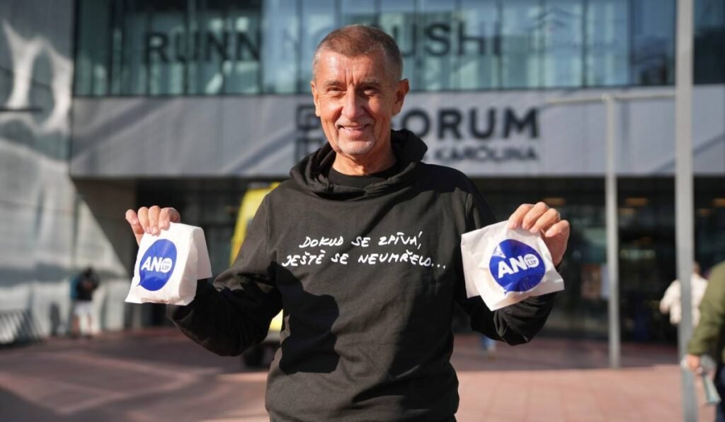 Miliardarul Andrei Babis. cu un hanorac negru cu un scris alb, cu mainile ridicate si cate o punguta alba cu inscriptie albastra in fiecare mana, a împărțit gogoși în prima zi de vot din Cehia, octombrie 2025