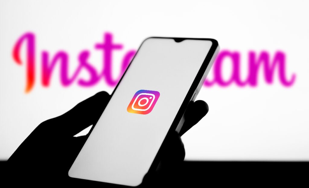 Imaginea arată silueta unei mâini care ține un smartphone pe al cărui ecran alb este afișată sigla aplicației Instagram – o pictogramă stilizată de cameră foto cu un gradient vibrant de culori. Pe fundal, estompat, se zărește logo-ul scris al rețelei sociale, subliniind focalizarea pe aplicația de pe telefon.