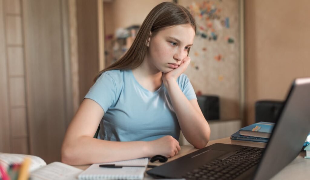 O tânără în tricou albastru deschis, care stă la un birou pe care se află un laptop și mai multe caiete