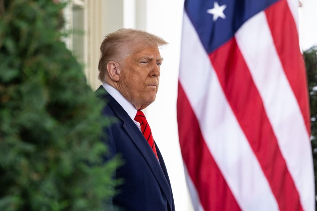 Donald Trump, in costum bleumarin si cravata rosie are in fata steagul SUA, iar in spate un copac verde